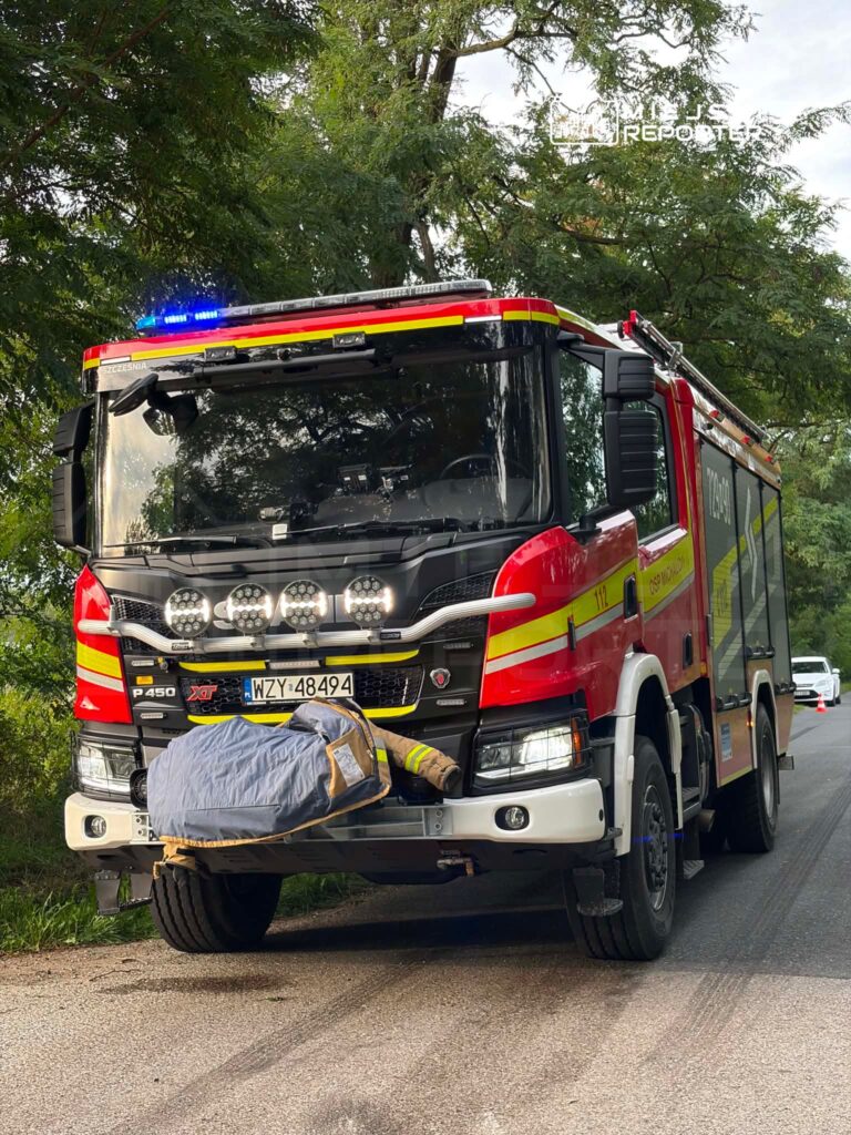 Czerwony wóz strażacki z niebieskimi światłami stoi na drodze, obok leży pokryty kocem strażak w mundurze.
