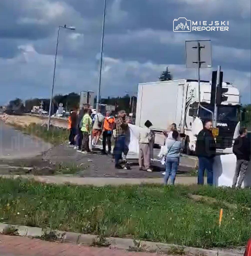 Grupa osób w kamizelkach odblaskowych trzyma transparenty na poboczu drogi, obok ciężarówki.