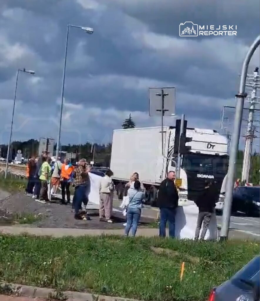 Grupa osób w odblaskowych kamizelkach i cywilnych ubraniach trzyma transparenty na poboczu drogi, obok przejeżdżających ciężarówek.