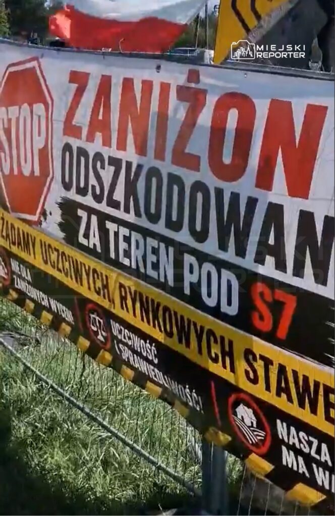 Transparent z napisem "STOP ZANIŻONĄ ODSZKODOWANĄ ZA TEREN POD S7" umieszczony na ogrodzeniu, w otoczeniu trawy i flagi.