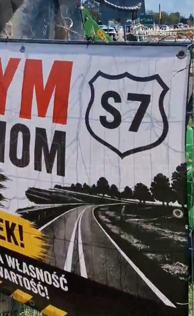 Transparent z napisem „S7” oraz hasłem „Zróbmy to razem!” w tle z widokiem na zakręt drogi otoczonej drzewami.