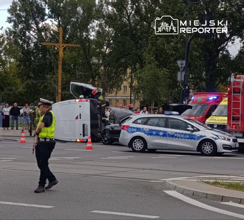 Na skrzyżowaniu stoi przewrócony pojazd, a obok zespół ratunkowy oraz policjant kierujący ruchem. W tle zgromadzeni świadkowie.