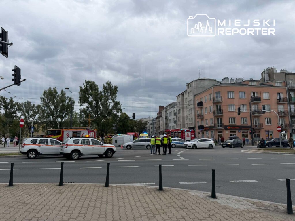 Grupa policjantów rozmawia na skrzyżowaniu, obok pojazdów ratunkowych i zaparkowanych samochodów. W tle widać budynki mieszkalne.