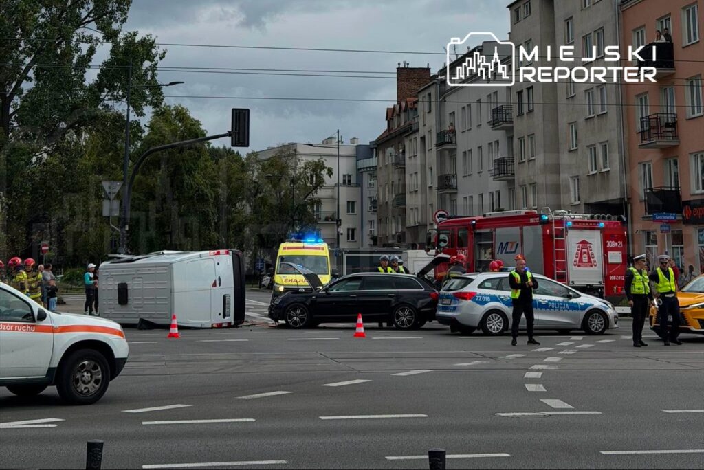 Na skrzyżowaniu stoi przewrócony biały furgon, obok niego zespół ratunkowy oraz policja zabezpieczająca miejsce zdarzenia.
