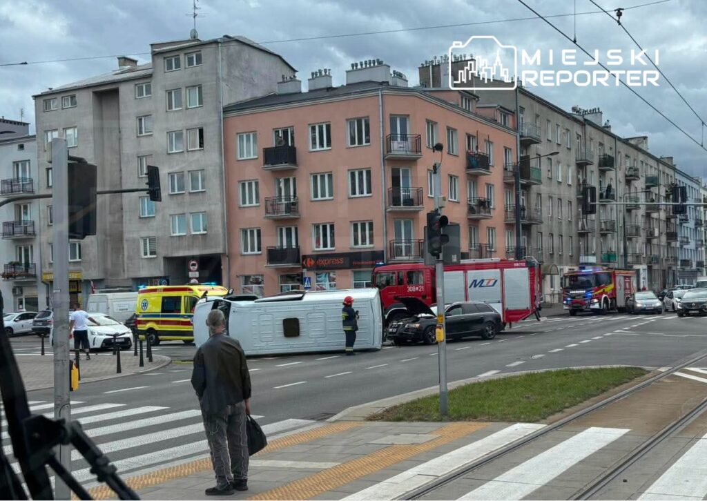 Na skrzyżowaniu ulic leży przewrócony pojazd, a obok niego stoją strażacy i karetka pogotowia. W tle widać budynki mieszkalne oraz przechodnia czekającego na zielone światło.