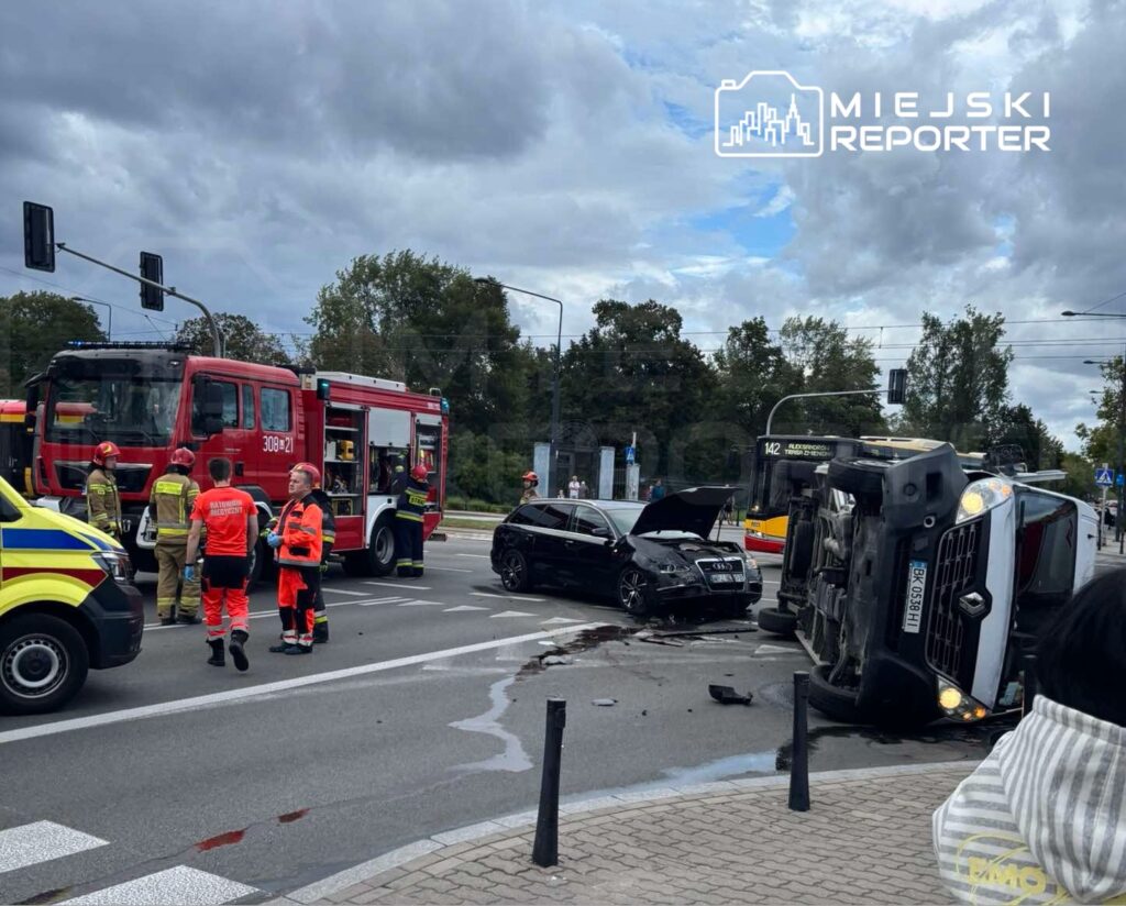 Na skrzyżowaniu leży przewrócony ciężarówka, obok stoi czarny samochód osobowy. Ratownicy i strażacy udzielają pomocy.