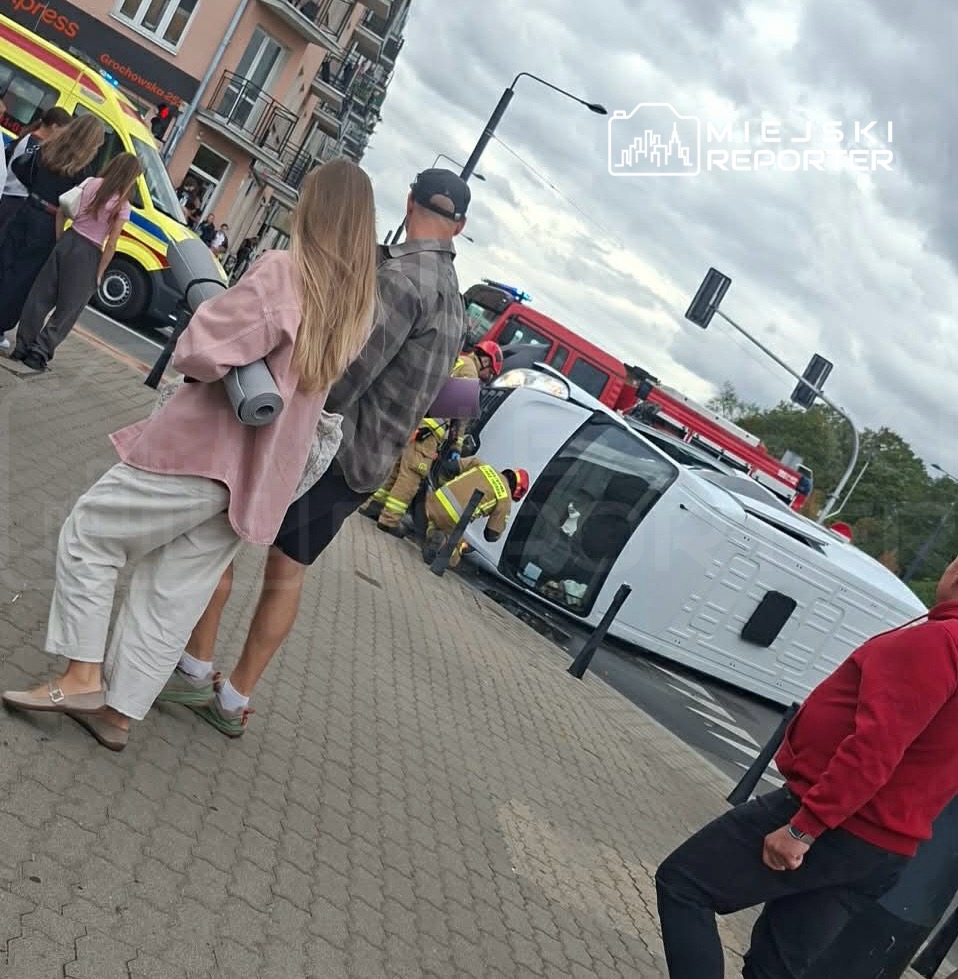 Kobieta z długimi włosami trzymająca matę, mężczyzna w czapce oraz grupa strażaków udzielających pomocy przy przewróconym samochodzie.