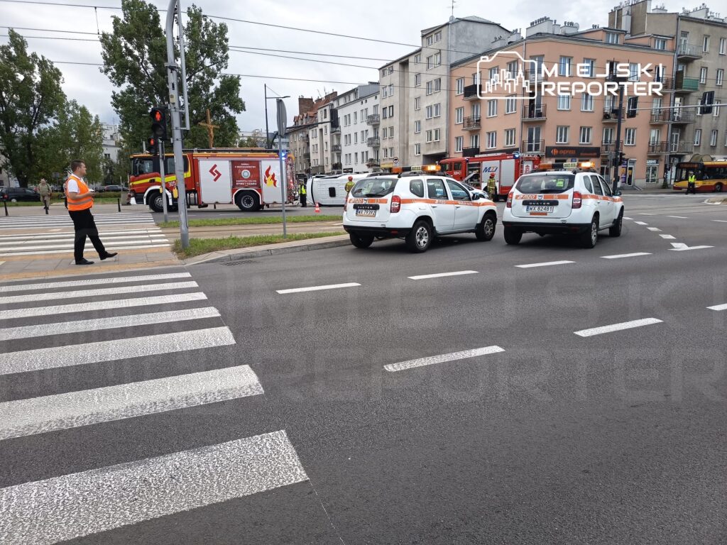 Mężczyzna w pomarańczowej kamizelce przechodzi przez przejście dla pieszych obok pojazdów straży pożarnej i policyjnych.