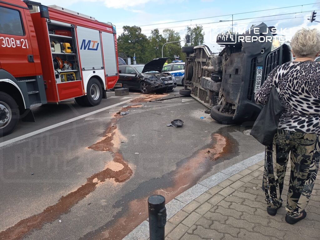 Na skrzyżowaniu leży przewrócony samochód, obok stoi wóz strażacki. Kobieta w wzorzystej bluzie obserwuje miejsce zdarzenia.