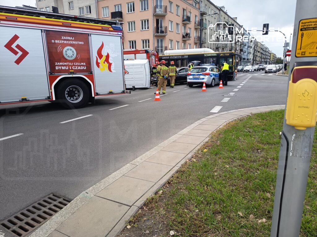 Strażacy i policjanci kontrolują miejsce zdarzenia drogowego, obok zaparkowany wóz strażacki i radiowóz.