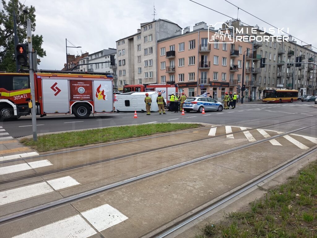 Na skrzyżowaniu ulic stoją strażacy przy wozie bojowym oraz policjanci, obok przewrócony pojazd.