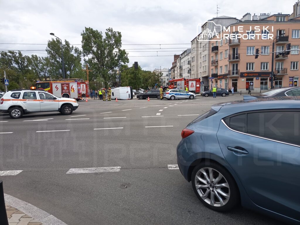 Na skrzyżowaniu stoją pojazdy straży pożarnej oraz policji, a w tle widoczne jest przewrócone auto. Przechodnie obserwują zdarzenie.