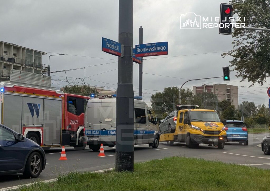 Na skrzyżowaniu ulic widać policyjny furgon oraz wóz strażacki z migającymi światłami. W tle holownik podnosi uszkodzone auto.