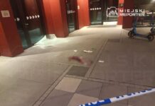 Rozbita szyba przy wejściu do budynku, na chodniku leżą porozrzucane papiery, otoczone taśmą policyjną.