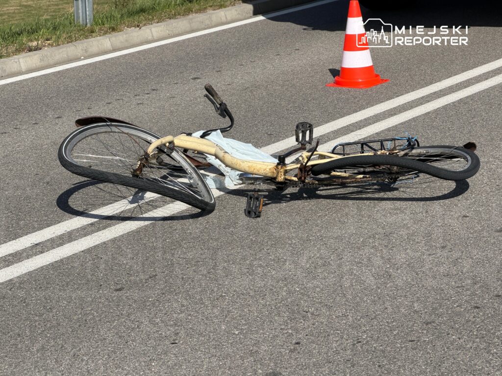 Rower leży na asfalcie, obok znajduje się stożek drogowy, w tle widać pasy ruchu.