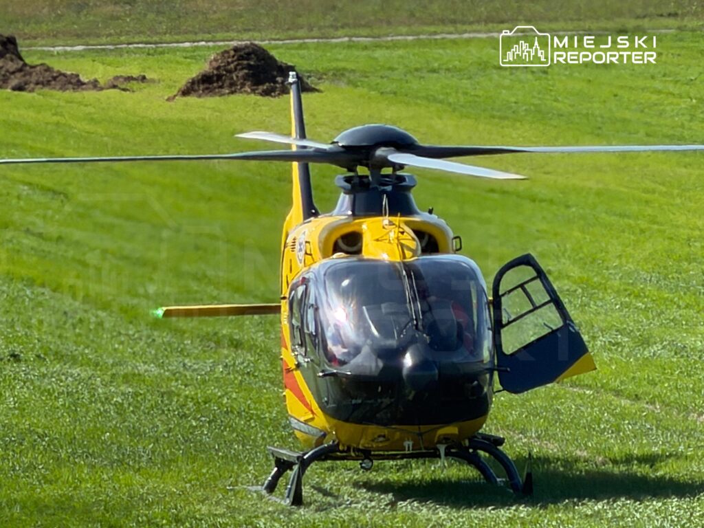 Żółty helikopter lądował na zielonym terenie, z otwartymi drzwiami i widocznymi wirnikami.