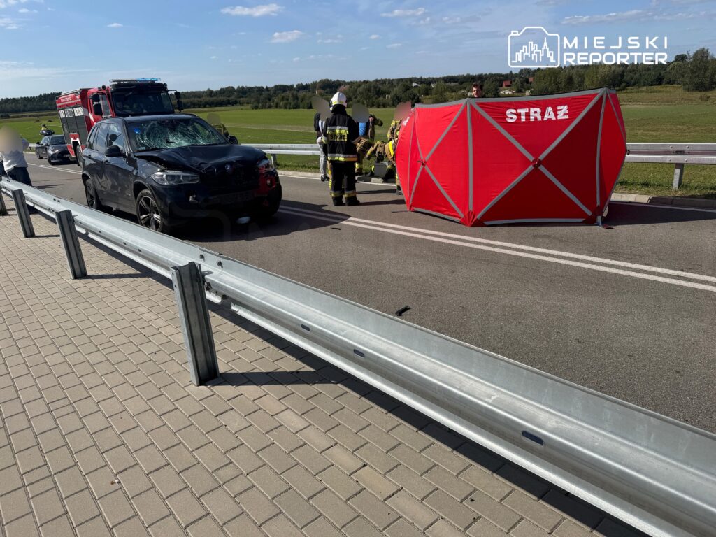 Czarny samochód osobowy z rozbitą szybą stoi na drodze obok czerwonego namiotu strażackiego, w tle zielone pola.