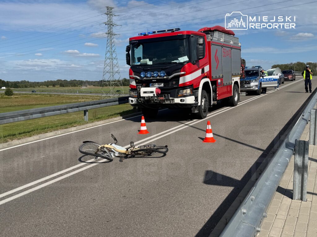Na drodze leży przewrócony rower, obok stoi wóz strażacki z włączonymi sygnałami świetlnymi.