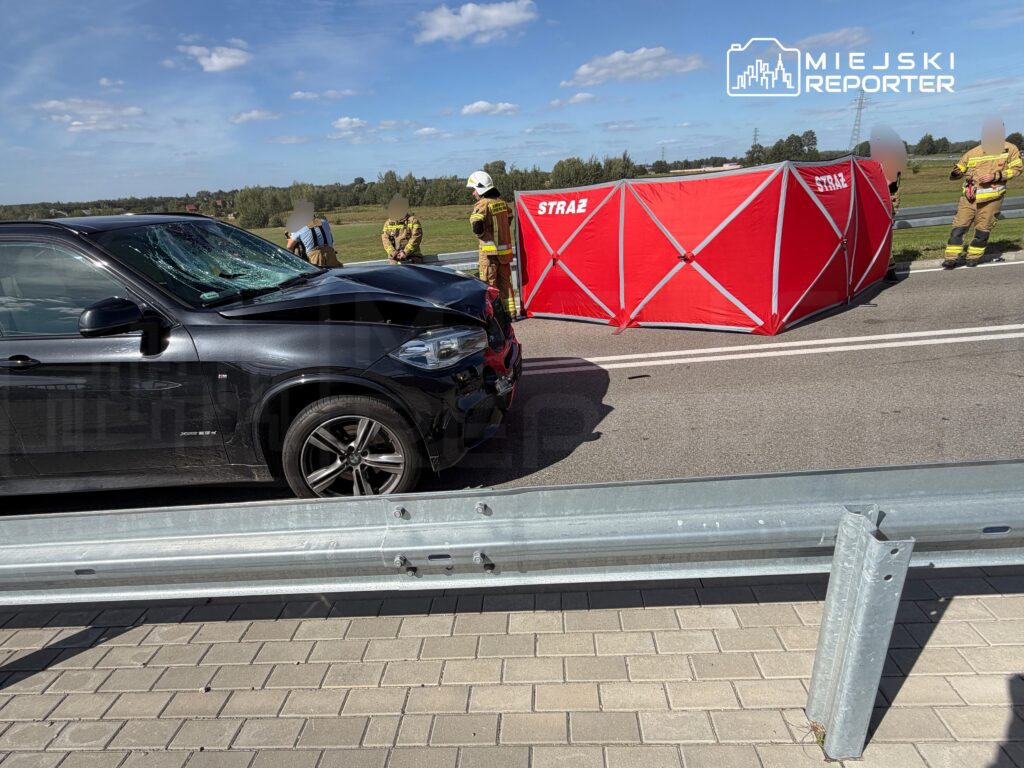 Czarny samochód osobowy z rozbitą szybą stoi na drodze, obok strażacy rozstawiają czerwony namiot.