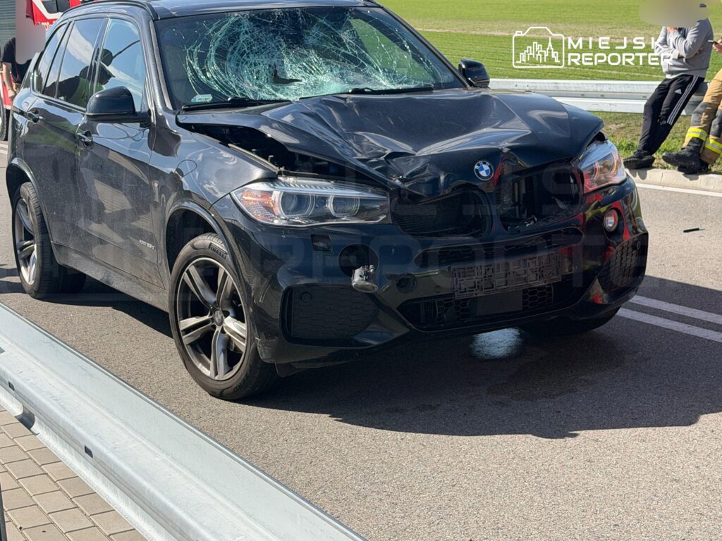 Czarny samochód marki BMW z uszkodzoną maską i zbitym przednim szybą stoi na drodze obok służb ratunkowych.