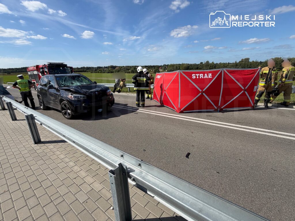 Na drodze stoi czarny samochód z uszkodzoną szybą, obok pracują strażacy i policjanci, a w tle widoczny jest czerwony namiot.