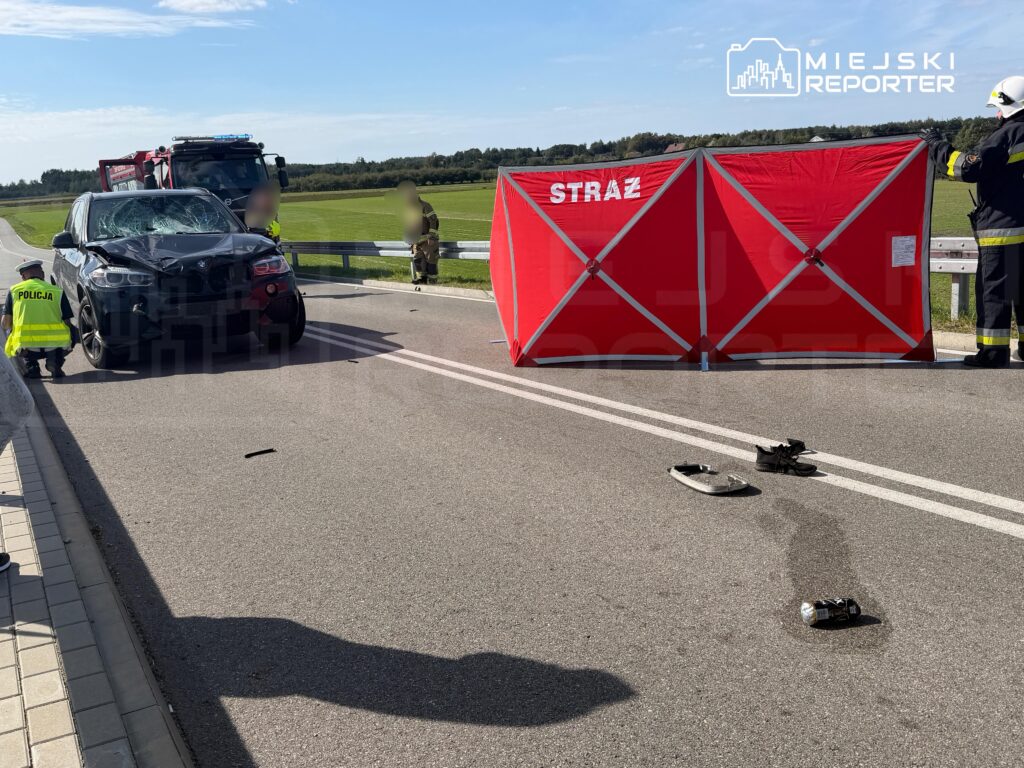 W miejscu wypadku drogowego stoi uszkodzony samochód osobowy, obok pracują policjanci i strażacy. Na jezdni leżą fragmenty obuwia i latarka.