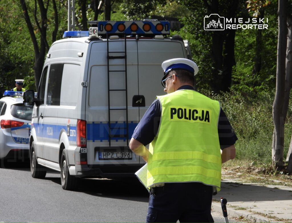 Policjant w kamizelce odblaskowej kontroluje dokumenty obok policyjnego samochodu na drodze.