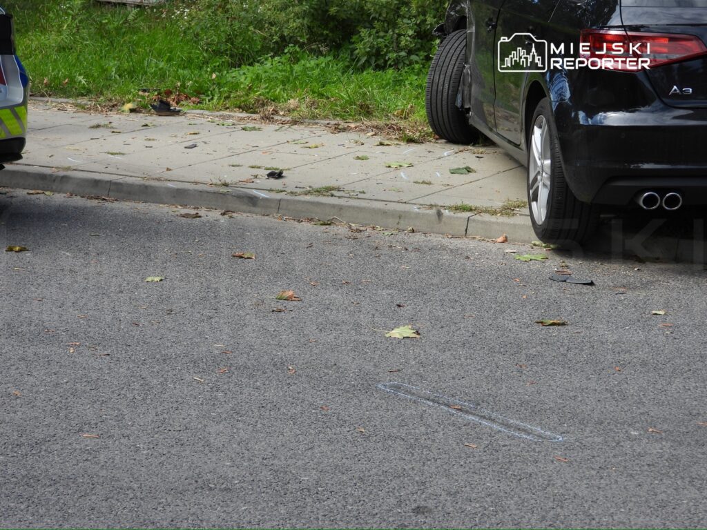 Czarny samochód osobowy zaparkowany na chodniku obok oznaczeń w białej kredzie, w otoczeniu liści na jezdni.