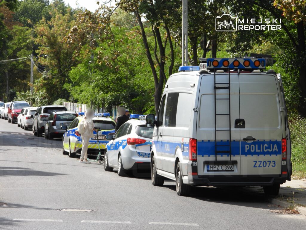 Policjant rozmawia z osobą w białym ubraniu obok radiowozu na ulicy otoczonej samochodami i drzewami.