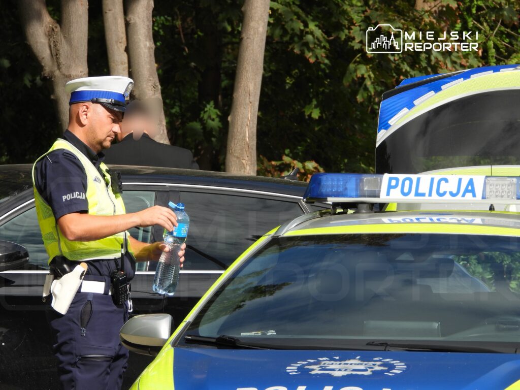 Policjant w kamizelce odblaskowej trzyma butelkę wody, stojąc obok radiowozu w otoczeniu drzew.