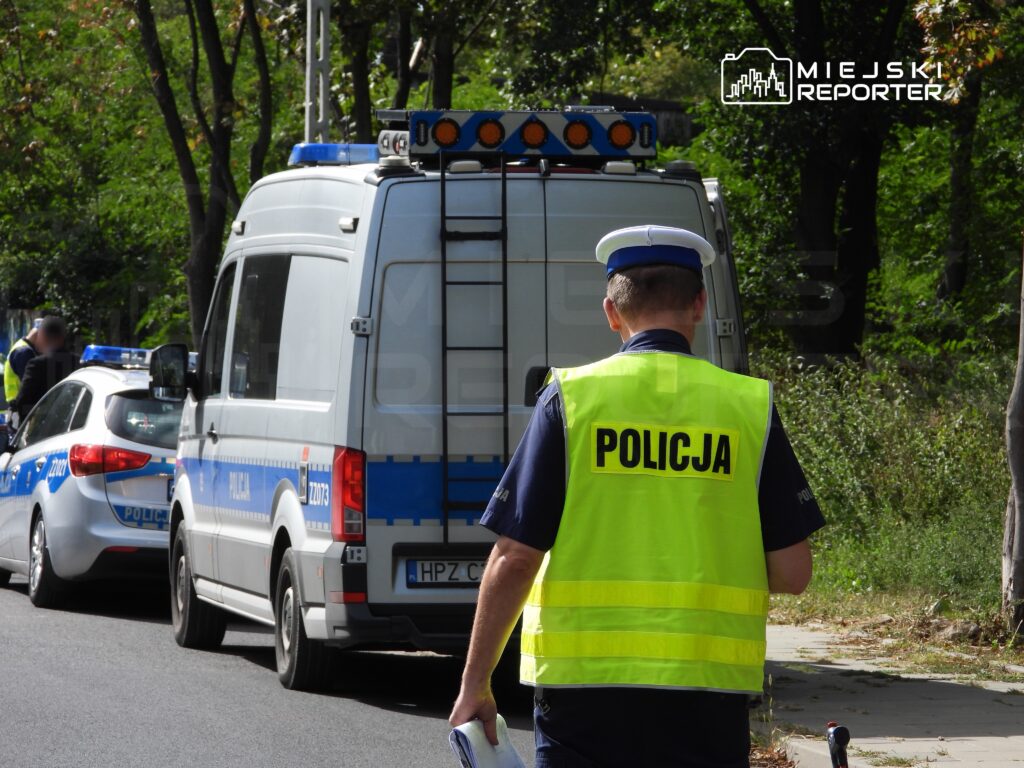 Policjant w kamizelce odblaskowej idzie wzdłuż ulicy obok radiowozu policyjnego i furgonetki z sygnalizacją.