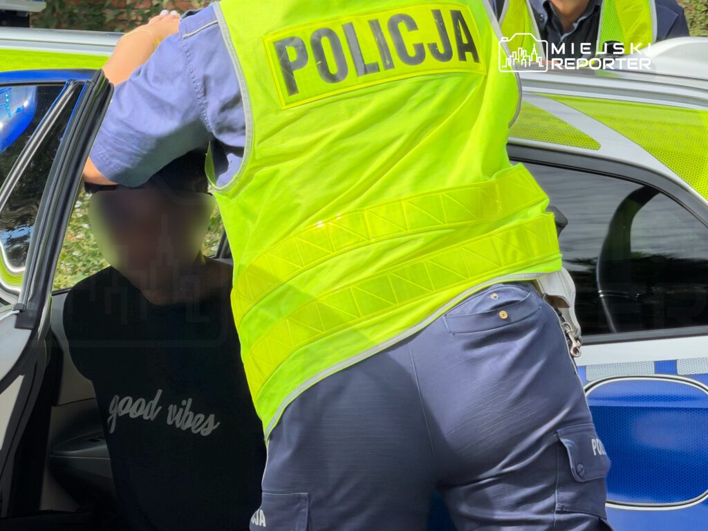 Policjant w odblaskowej kamizelce zatrzymuje osobę siedzącą w samochodzie osobowym.