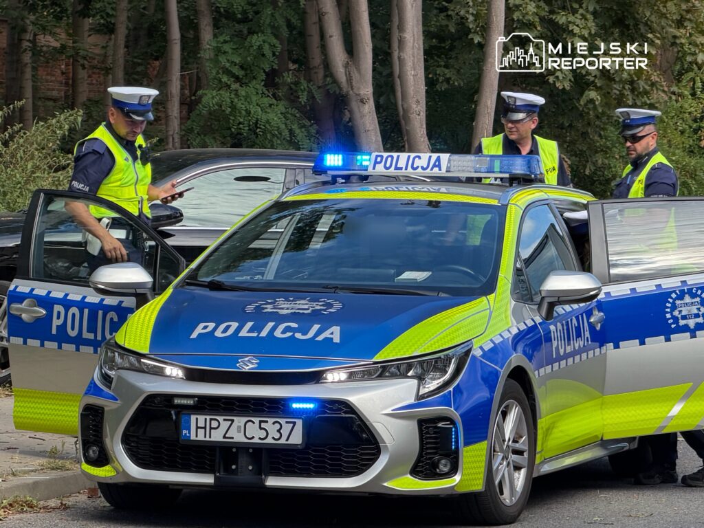 Policjanci w mundurach interweniują przy radiowozie, sprawdzając dokumenty w leśnym otoczeniu.