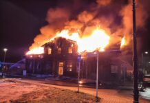 Płonący budynek z drewnianą fasadą, ogień wydobywa się z dachu, a strażak polewa go wodą z węża.