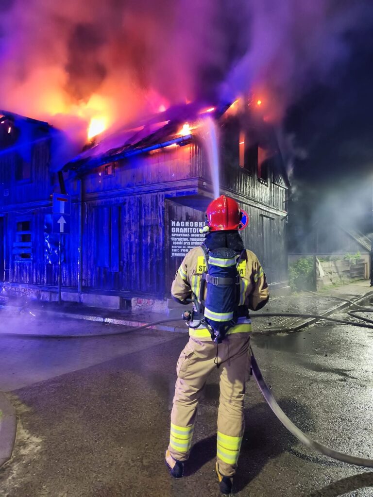 Strażak w hełmie gasi pożar drewnianego budynku, z którego wydobywają się płomienie i dym w nocnym otoczeniu.