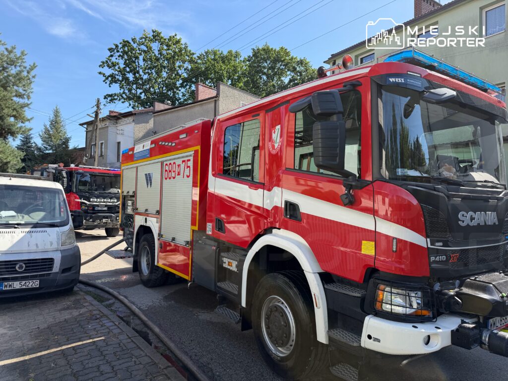 Wóz strażacki marki Scania z włączonymi sygnalizacją świetlną stoi na ulicy obok białego furgonu, w tle widoczne budynki.