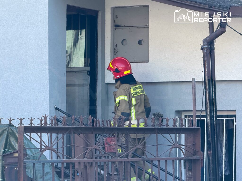 Strażak w hełmie i mundurze wchodzi do budynku przez otwarte drzwi, w tle widoczny metalowy płot.