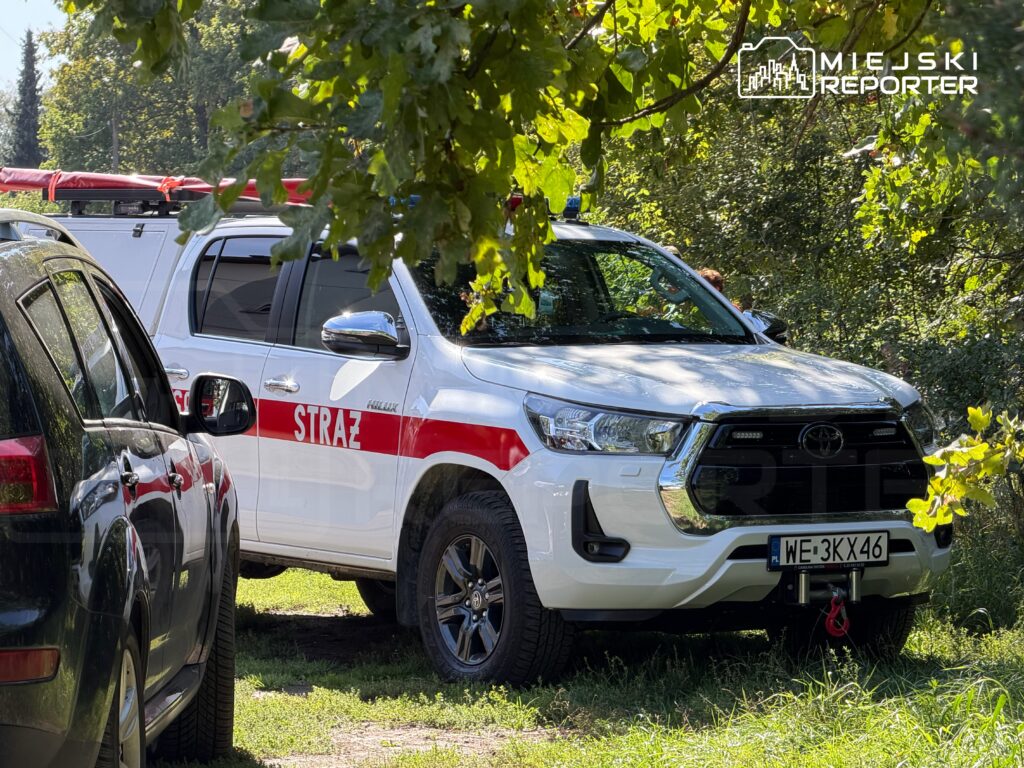 Samochód straży pożarnej z napisem "STRAŻ" zaparkowany w zielonym otoczeniu, obok innych pojazdów.