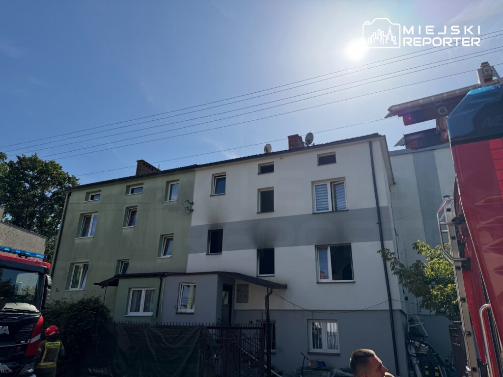 Czerwony wóz strażacki stoi przed budynkiem mieszkalnym z otwartymi oknami, obok strażak rozmawia z mężczyzną.