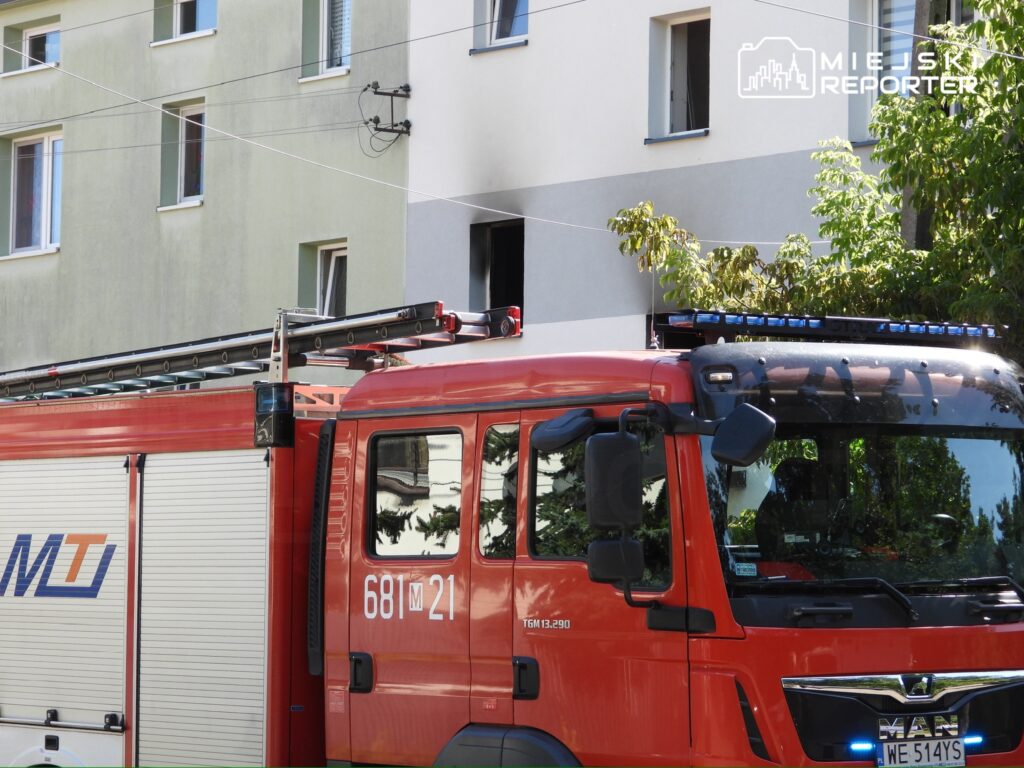 Czerwony wóz strażacki z widoczną drabiną stoi przed budynkiem mieszkalnym w otoczeniu drzew.