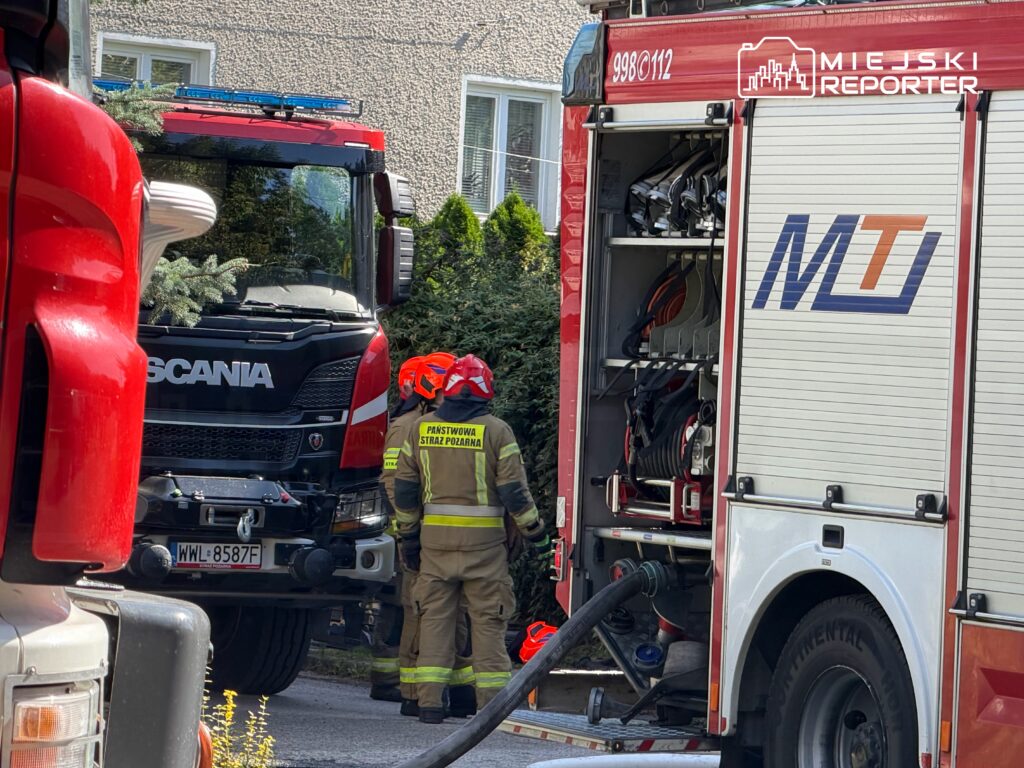 Strażacy w hełmach rozmawiają przy wozie strażackim Scania, obok drugiego wozu z otwartą klapą.