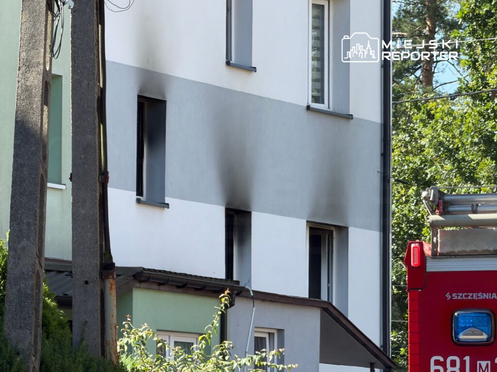 Czarna smuga na ścianie budynku wskazuje na uszkodzenie po pożarze, obok stoi wóz strażacki.