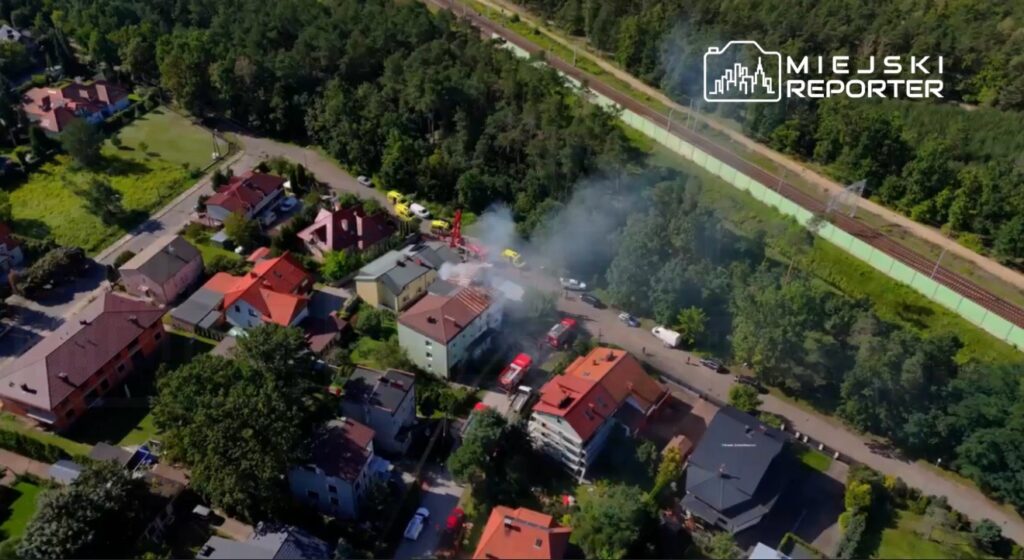 Dym unoszący się z budynku mieszkalnego w zabudowie jednorodzinnej, otoczonym drzewami i drogami. W tle widoczne pojazdy straży pożarnej.
