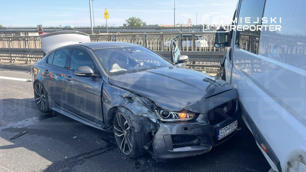 Uszkodzony samochód osobowy stojący na drodze, z widocznymi uszkodzeniami przodu i boku, obok dużego pojazdu dostawczego.