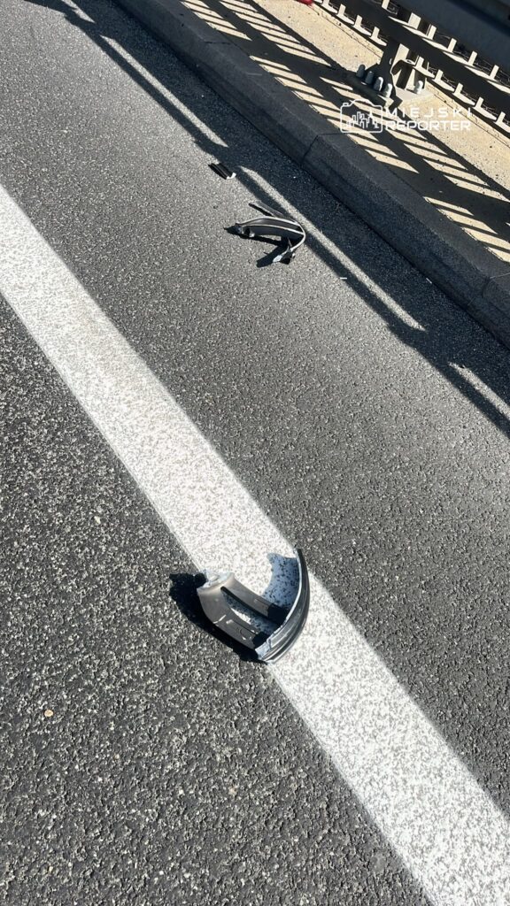 Na drodze leży uszkodzona część pojazdu obok białej linii. W tle widoczny jest metalowy barierka.