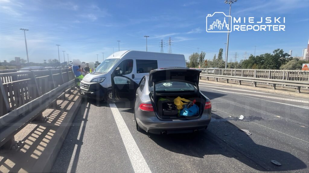 Na drodze stoi szary samochód osobowy z otwartym bagażnikiem, obok niego zaparkowany biały furgon. Dwóch mężczyzn w kamizelkach odblaskowych rozmawia obok pojazdu.