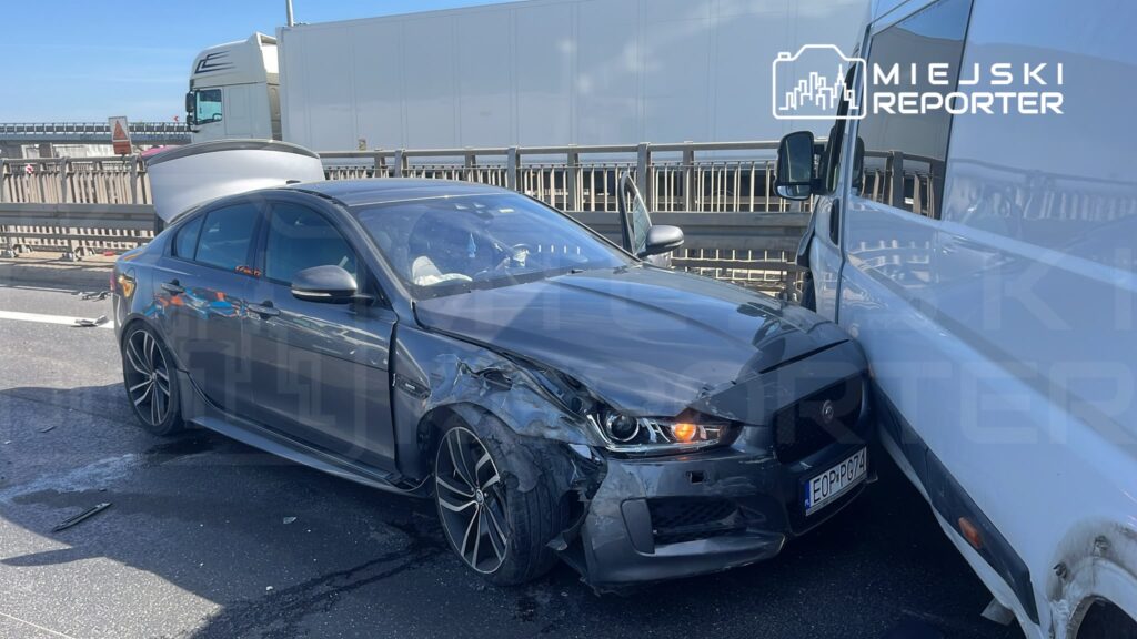 Czarny samochód osobowy z uszkodzoną przednią częścią stoi na drodze, obok białego pojazdu dostawczego.