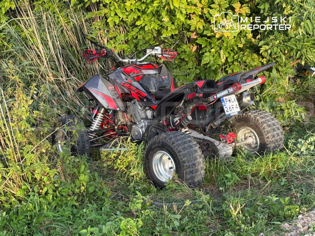 Czarny quad z czerwonymi akcentami stoi wśród wysokiej trawy i krzewów.