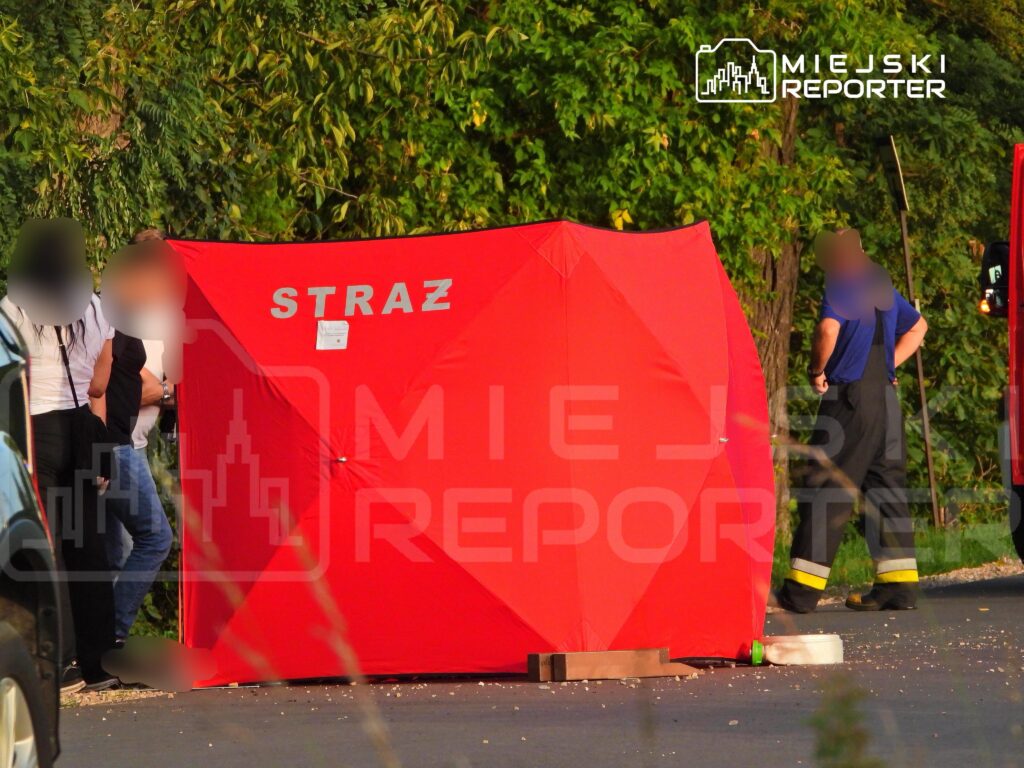 Grupa osób stoi obok czerwonego namiotu z napisem "STRAŻ" w otoczeniu zieleni, w pobliżu drogi.