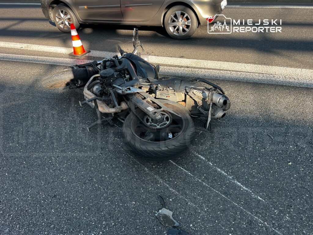 Motocykl leżący na drodze, obok czerwono-biały słupek ostrzegawczy, przyjeżdżające samochody w tle.